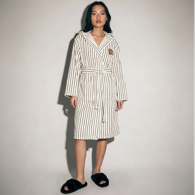 A model stands against a plain light gray wall wearing a cream and black vertically striped hooded robe with a tie belt, black fuzzy slippers, and a small embroidered crest on the chest.